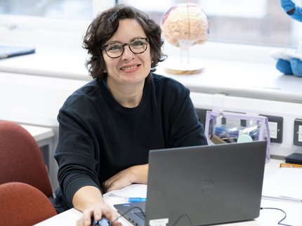 A middle aged woman with wavy dark hair and glasses sits at a computer desk smiling.