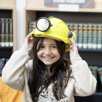 A young girl smiling in a library with a miners helmet on