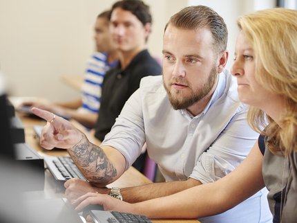 White female adult learner with blonde mid length hair wearing a grey sleeveless top sits at a computer with a white male learner with tattoos, short hair and a beard.
