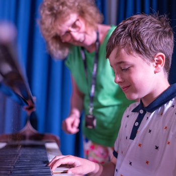 A woman in a green top points to a music sheet on the piano with a child playing the keys
