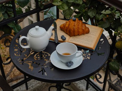 A photograph of an open book on a table. Also on the table is a croissant and some bluberries