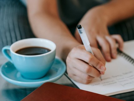 A pen is poised in a hand ready to write a poem in a notebook beside a hot drink.