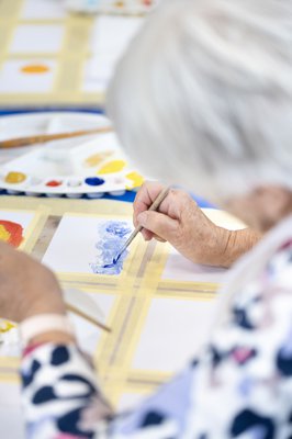 An older person's hands using paint brushes, surrounded by tubes of paint and an artist's palette.