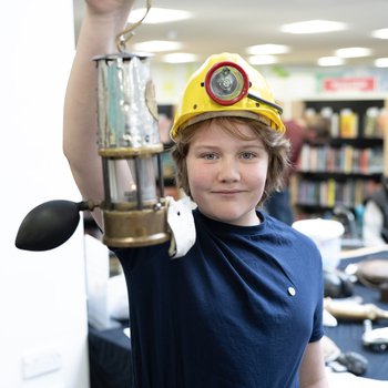 Child wearing a miners helmet and holding up an oil lamp smiling