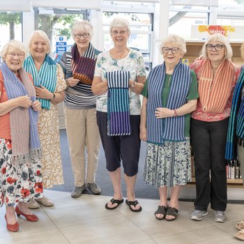 A group of elderly ladies standing in a row holding knitted scarves