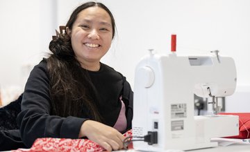 Smiling learner with dark hair wearing a black long sleeve t-shirt sits at a sewing machine using red patterned fabric and red cotton.