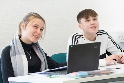 Two students sat at a desk, one has a laptop open in front of her.