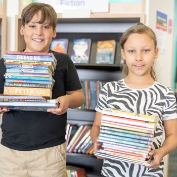 a boy and girl in a library smiling, each holding a pile of books.