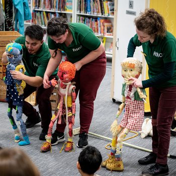 Three actor/puppeteers from the show What Happened to You? taking place in a library hold and stand behind three raggedy puppets, Bo, Little Red and Noodle.